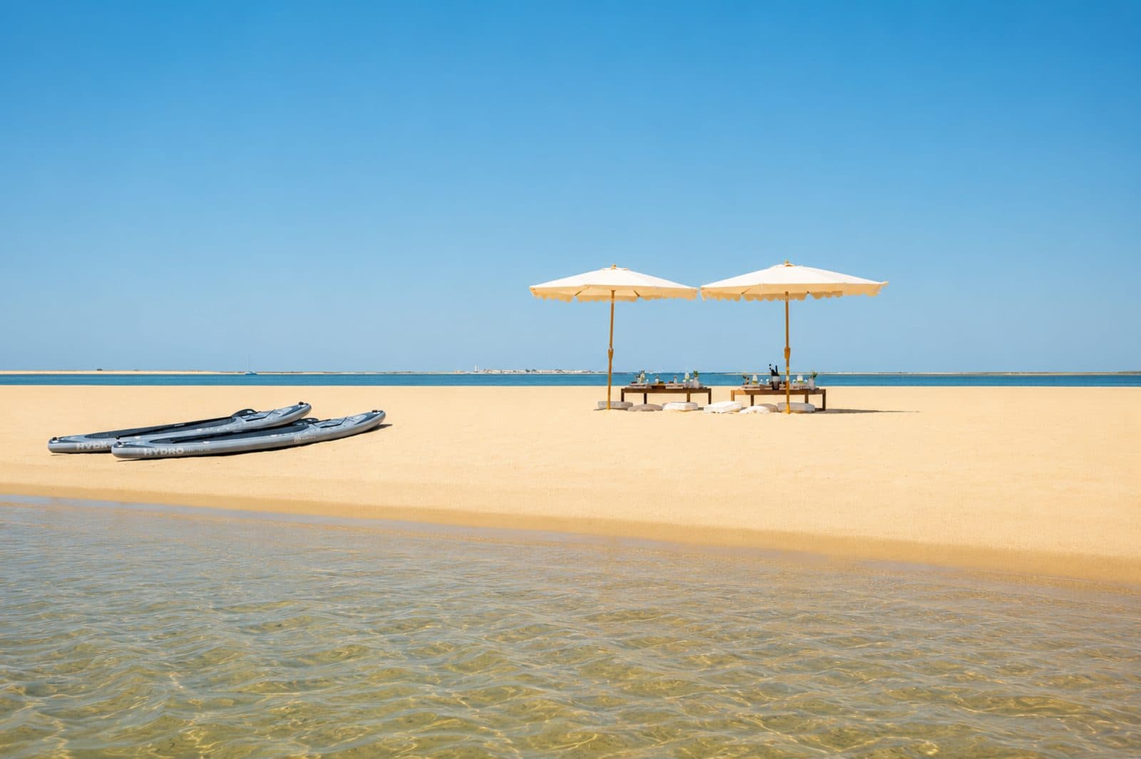 Two parasols and paddle boards on the sand beside clear shallow water